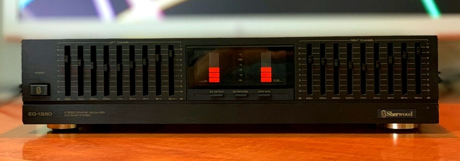 A stock image of a Sherwood EQ-1330, showing the same positioning of the EQ sliders and VU meters as in the screenshot from The Matrix. It's a black unit, with orange VU meters, the VU meters are segmented into rectangles, and the topmost segment on both left and right VU meters is red instead of orange. There are several EQ band sliders for left and right channels, to the left and right of the VU meters. They are labelled with the various cutoff frequencies between 63Hz and 16Khz.